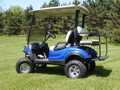 Blue golf cart with off-road tires parked on grass in front of a tree line.