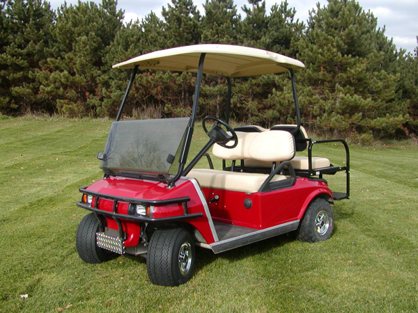 A red golf cart with a beige roof and seats is parked on a grassy area with pine trees in the background.